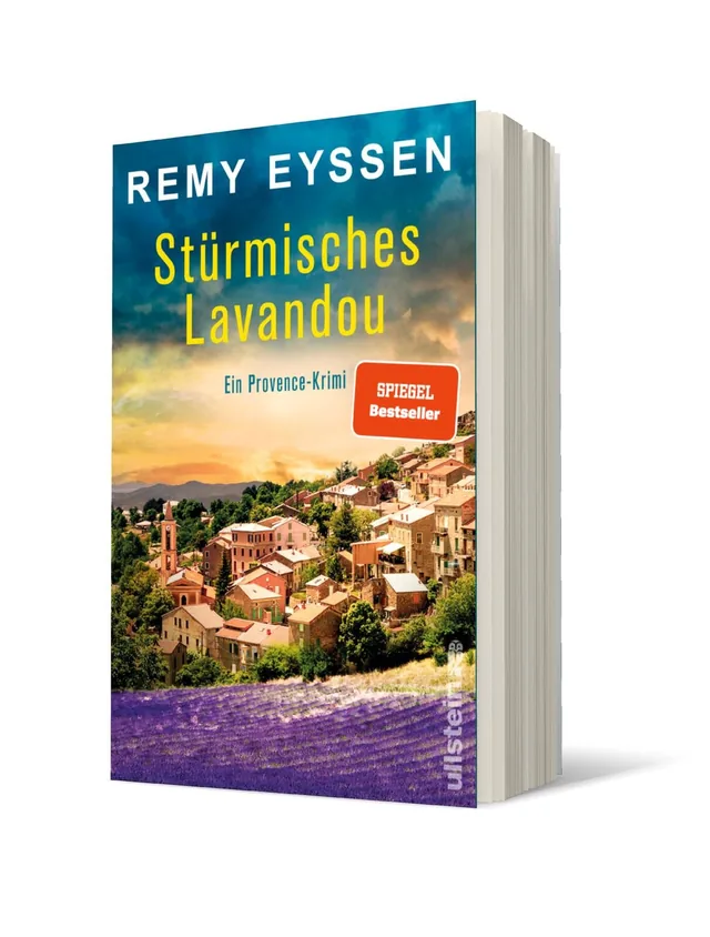 Cover von „Stürmisches Lavandou“ von Remy Eyssen: Die Abbildung zeigt eine malerische Provence-Landschaft mit bunten Gebäuden, grünen Hügeln und lila Lavendelfeldern unter einem blauen Himmel.